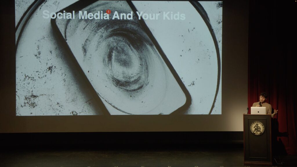 A person standing at a podium in front of a large screen. The screen features an image of a cell phone and the text "Social Media And Your Kids"